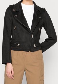 Veste motard noire en suédine synthétique avec une coupe ajustée, poches zippées et fermeture à pression au col. Portée sur une chemise blanche.