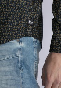 Camisa de patrón en azul oscuro y diseño geométrico dorado, con una etiqueta tejida, combinada con unos pantalones de mezclilla azul claro con una textura desgastada.