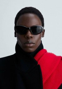 Zwarte zonnebril met een slank gebogen frame, gedragen met een zwarte jas met een rode ribbelaccent, wat een gedurfde contrast in texturen geeft.