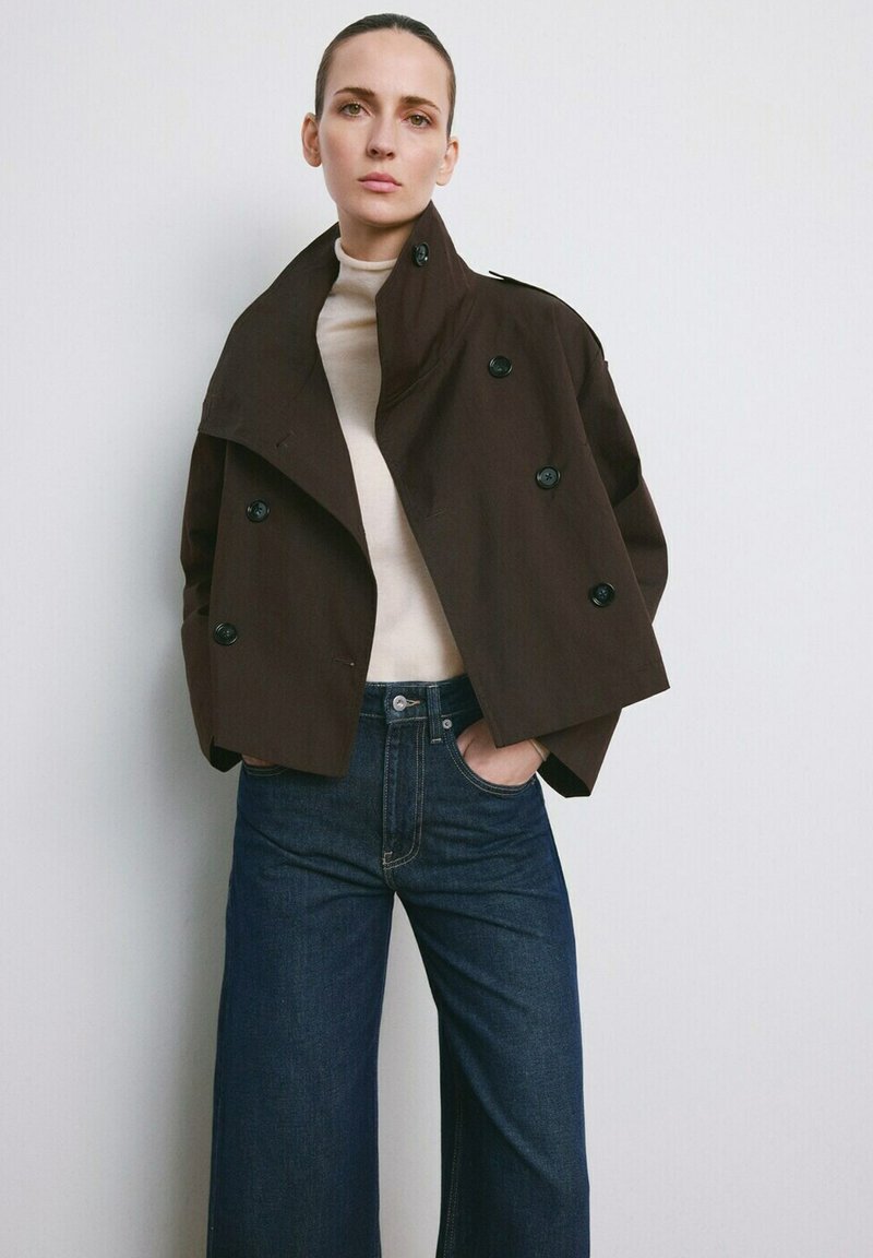 Ženska v temno rjavi kratki dvoredni jakni, bež pajkici z visokim ovratnikom in visokim pasom širokih temno modrih jeans hlač, stoji ob enobarvni steni.