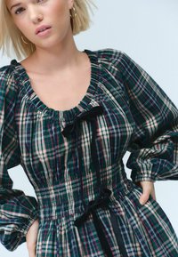 Geruit jurkje in groen, marineblauw en beige met een gathered halslijn, pofmouwen en zwarte lintaccenten in de taille. Gerimpelde taille detail.