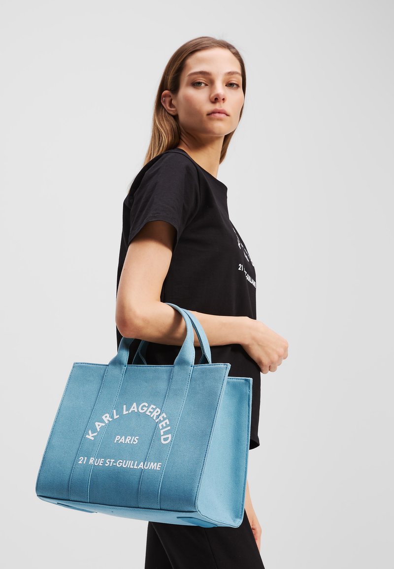 Borsa a spalla azzurro chiaro con tessuto testurizzato, caratterizzata da un logo e testo stampati bianchi. Manici corti per il trasporto. Il modello indossa una maglietta nera.