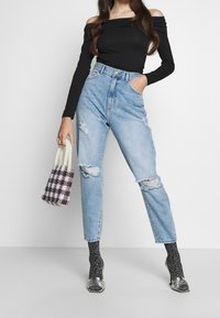 Kvinna i svart avskuret topp, högmidjade ljusblå jeans med hål, mönstrade strumpor med metalliska öppen-tå klackar, håller en pärlhandväska.