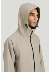 Mand iført beige regnjakke med hætte, sort lynlås og justerbare snørelåse, kigger mod højre mod en lysegrå baggrund.