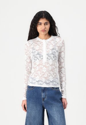 Long-sleeve top in sheer white fabric with floral lace pattern, round neckline, and button closure. Paired with high-waisted denim jeans.