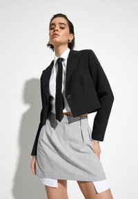 Blazer noir cropped sur une chemise blanche avec une cravate noire, associé à une mini jupe plissée grise. Textures lisses et lignes nettes.