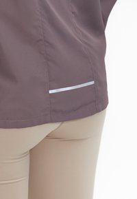 Lilla, letvægtsjakke med en glat overflade, der har en sølvreflekterende stribe nær kanten. Beige tætsiddende bukser er synlige nedenunder.