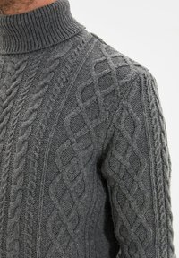 Grauer Strickpullover mit hohem Rippenkragen und aufwendigen Zopfmustern über dem Körper. Das strukturierte Material sorgt für Wärme.