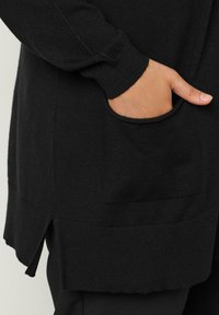 Schwarze, gestrickte Strickjacke mit langen Ärmeln, einer Tasche und gerippten Bündchen. Der Saum hat seitliche Schlitzöffnungen für mehr Bewegungsfreiheit. Weiche Textur.