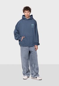 Sudadera azul de gran tamaño con capucha grande, bolsillo frontal y gráficos impresos. Combinada con pantalones vaqueros de pierna ancha de color azul claro con patrones de llamas.