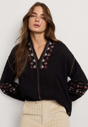 Frau mit langen braunen Haaren, die eine schwarze Bluse mit roter und weißer geometrischer Stickerei und beige gerippte hochgeschnittene Hose trägt.