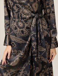 Rochie neagră cu modele paisley și florale aurii elaborate, având talie legată și mâneci lungi dintr-un material neted și texturat.