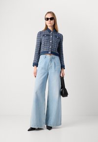 Veste bleu cropped avec un motif texturé et des boutons, assortie à un jean large bleu clair. Le mannequin tient un petit sac à main noir.