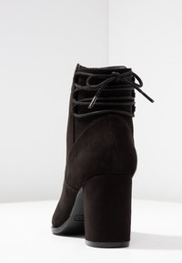 Stivale alla caviglia in suede nero con tacco a blocco, dettaglio con lacci sul retro e punta tonda chiusa, su sfondo bianco.
