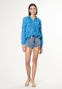 Blauwe chiffon blouse met ruches, gecombineerd met versleten denim short. Model draagt witte sandalen met banden. Eenvoudige achtergrond.