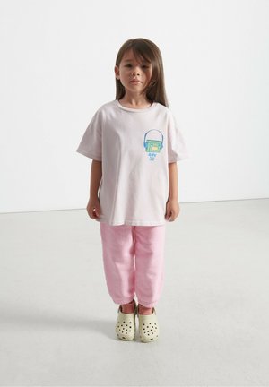 Lichtroze katoenen T-shirt met een grafische print, gecombineerd met roze joggingsbroeken en gele klompen. Eenvoudig ontwerp, relaxed fit en comfortabele textuur.
