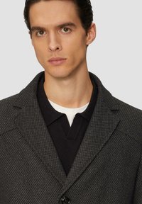 Dunkelgrauer strukturierter Blazer mit breiten Revers, Ein-Knopf-Verschluss und einem schwarzen Poloshirt darunter. Klare Linien und taillierte Passform.
