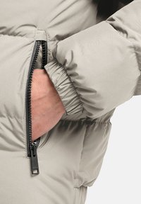 Manica di giacca a vento in tessuto grigio chiaro con tasca con zip nera. L'orlo della manica è elastico, con una texture liscia e opaca. Dettaglio della zip visibile.