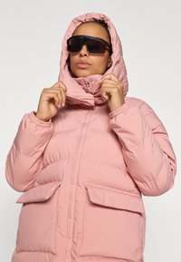 Roosa puffjakker kõrge krae ja kapuutsiga. Omab elastseid mansette ja kahte esimest patch taskut. Sile kangas õmblusdetailidega.