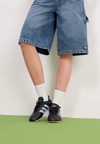 Denim šorti ar brīvu piegriezumu, zila krāsa, ar salocītām malām. Melni sporta apavi ar ziliem svītrainiem, apvienoti ar baltām cepures zeķēm. Zaļa grīdas fona.