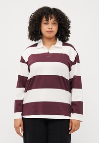 Une personne avec des cheveux bouclés porte une chemise polo à manches longues rayée bordeaux et blanche et se tient devant un fond uni.