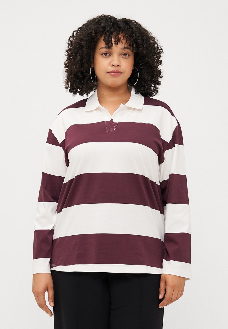 Une personne avec des cheveux bouclés porte une chemise polo à manches longues rayée bordeaux et blanche et se tient devant un fond uni.