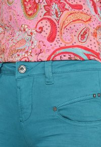 Tyrkysové džíny s hladkou texturou, s popínacím knoflíkem ve stříbrné barvě, postranními kapsami a dekorativními cvočky na kapse. Nad nimi růžová blůza s paisley vzorem.