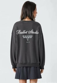 Siv svetrov z napisom "Baletna šola" v belem kurzivnem tisku na hrbtni strani. Ima sproščen kroj ter rebraste manšete in rob. Uporabljen z temno krilom.