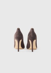 Scarpe con tacco alto in suede marrone scuro, con punta affilata e tacco a stiletto sottile. Presentano dettagli con logo dorato sul retro.