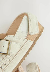 Sandale en daim blanc avec un design tissé, boucle réglable, semelle texturée marron et doublure intérieure douce et lisse.