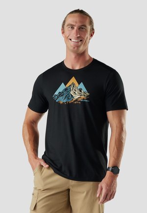 Uśmiechnięty mężczyzna z długimi włosami nosi czarną koszulkę z grafiką gór oraz khaki krótkie spodnie typu cargo, stoi z jedną ręką w kieszeni.