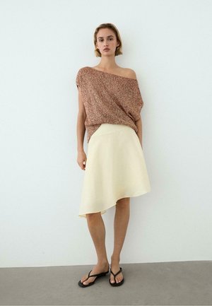 Femme portant un top marron texturé à une épaule, une jupe asymétrique crème et des sandales noires, debout contre un mur blanc uni.