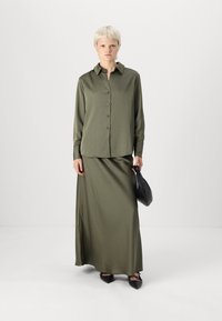 Chemise olive à manches longues avec des boutons et jupe maxi assortie, accompagnée d'un sac à main noir et de ballerines pointues, sur un fond neutre.