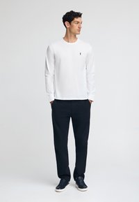 Maglietta bianca a maniche lunghe con un piccolo logo sul petto, abbinata a pantaloni scuri e sneakers blu navy. Tessuto morbido, vestibilità rilassata.