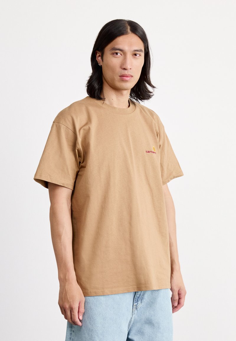 Brown cotton t-shirt with a crew neck and short sleeves. Features a small red logo on the chest and a relaxed fit. Light blue denim pants.