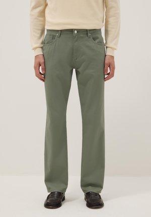 Hombre con pantalones rectos de color verde oliva, suéter crema y mocasines negros, de pie con los brazos relajados a los lados frente a un fondo liso.