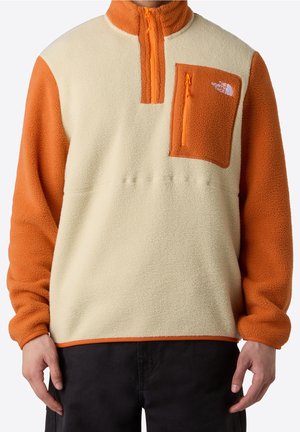 Mann trägt einen beigen und orangen Fleece-Pullover mit einer Reißverschlusstasche auf der Brust und dem Logo von The North Face.