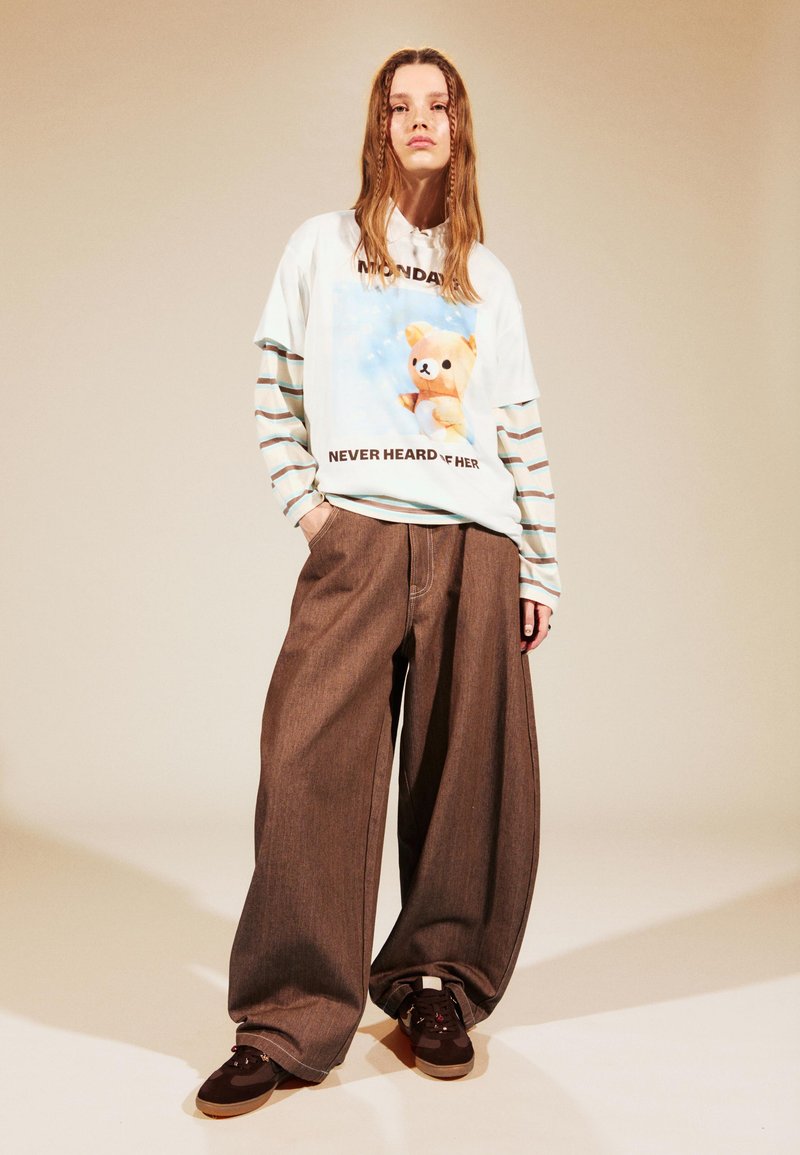 Biely tričko s grafickým motívom karikatúrneho medveďa, vrstvené cez pruhované tričko s dlhým rukávom. Široké hnedé nohavice a tmavé topánky dotvárajú outfit.