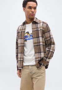 Νεαρός άνδρας που φοράει πουκάμισο με καρό σε μπεζ και καφέ αποχρώσεις πάνω από λευκή μπλούζα με γραφικά ψαριών και μπεζ παντελόνι, στέκεται με το ένα χέρι στην τσέπη.