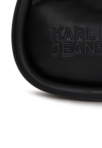 Crna kožna torbica s utisnutim logotipom "KARL JEANS", okruglog oblika, glatke teksture, minimalističkog dizajna i bez vidljivih metalnih dijelova.