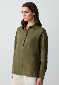Camicia in lino verde oliva con collare button-down, maniche lunghe e vestibilità morbida, caratterizzata da bottoni in tinta e superficie texturizzata.