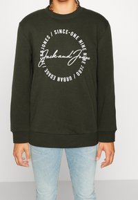 Felpa verde scuro realizzata in misto cotone, con testo bianco e logo in un design circolare. Maniche e orlo a costine.