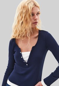 Blonde Frau mit lockigem Haar, die ein dunkelblaues Langarmoberteil über einem weißen gerippten Inner-T-Shirt trägt und nach rechts vor einem grauen Hintergrund schaut.