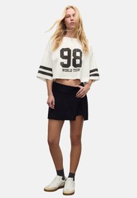 Witte cropped sweatshirt met zwarte "98 WORLD TOUR" opdruk en strepen, gecombineerd met een zwarte wikkelskirt en witte sneakers.