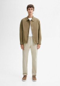 Olive green, lightweight jacket with a collar and snap buttons, worn over a white shirt. Beige pants and brown shoes with laces complete the look.