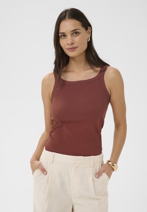 Mujer con cabello largo y oscuro, vistiendo una blusa sin mangas marrón, pantalones crema, pendientes de aro dorados y una pulsera de oro, con las manos en los bolsillos.