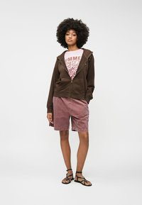 Pruun vetthoolik roosa graafilise T-särgi peal, koos heleda punase sametist lühikesed püksid ja mustade rihmadega sandaalidega. Inimene seisab valge tausta vastu.