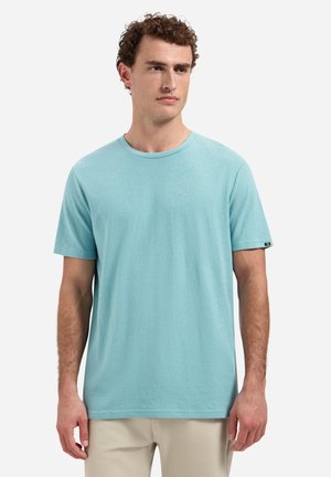 Man wearing plain light blue short-sleeve t-shirt and beige pants, standing and looking slightly to the side against a white background.