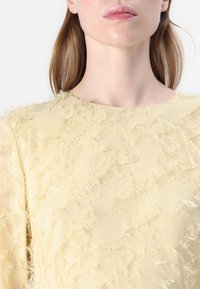 Gelbes, strukturiertes Oberteil mit ausgefransten Stoffdetails, Rundhalsausschnitt und transparenten langen Ärmeln, das einen leichten, gestuften Look betont.