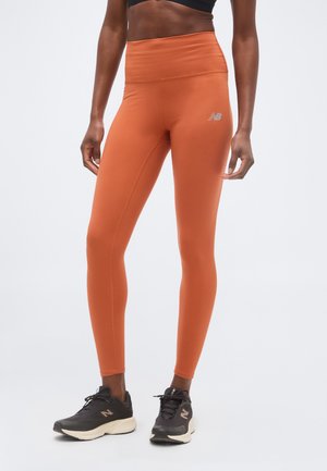 Femme portant des leggings taille haute orange rouille et des chaussures de sport noires, debout devant un fond uni.